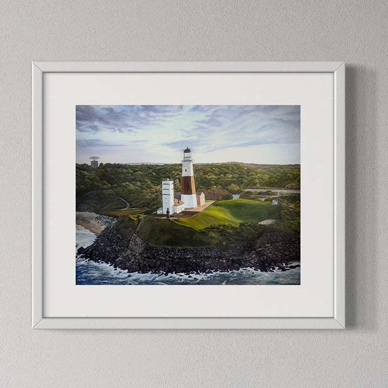 Framed artwork of a lighthouse on a rocky island with a body of water and green landscape.