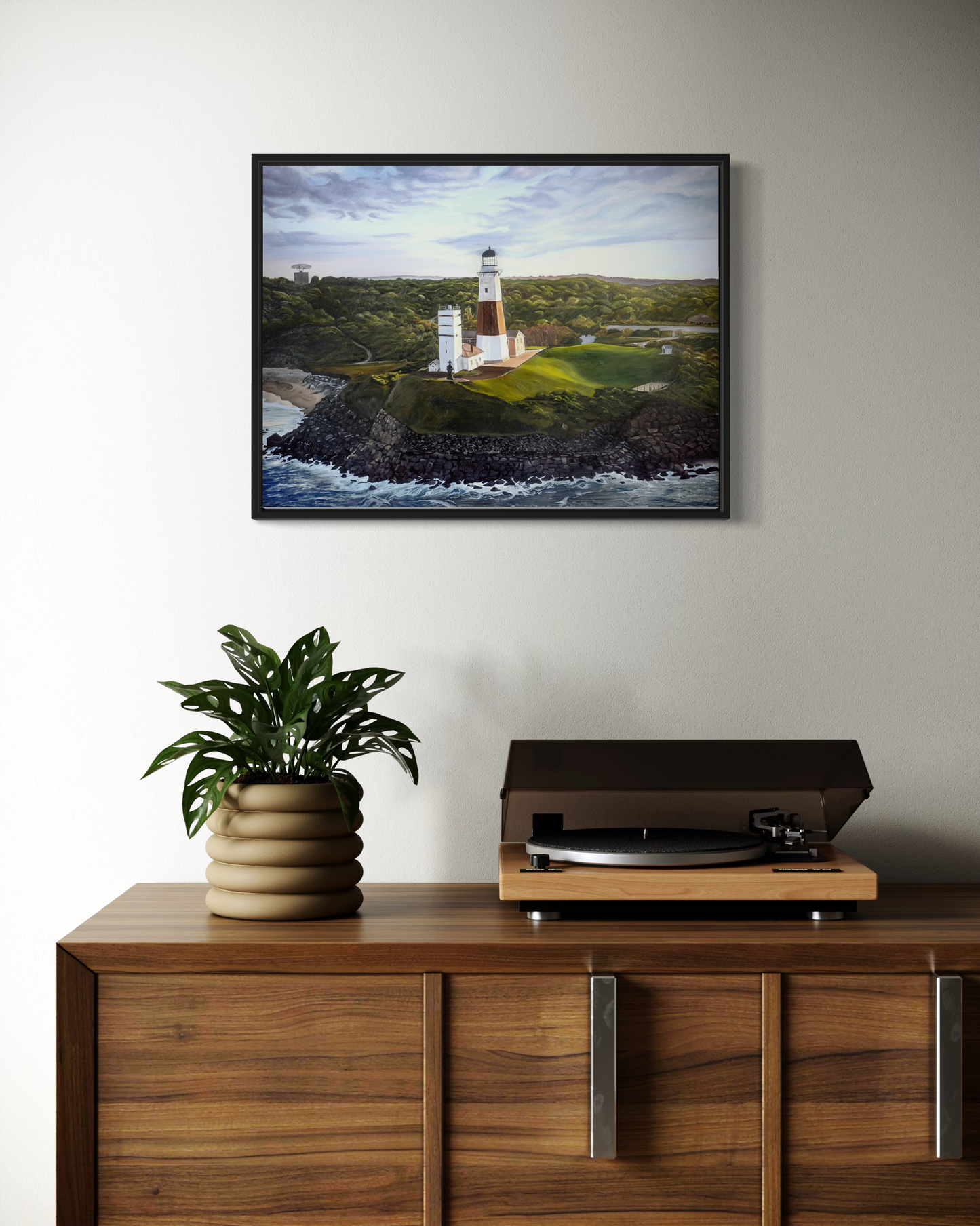Framed artwork of a lighthouse on a wall above a wooden cabinet with a plant and record player.