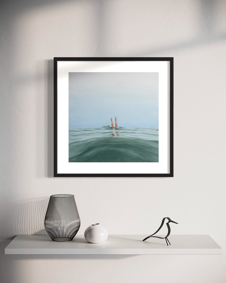 A framed print of 'Double Dipped, I' hanging on a wall above a shelf with a decorative vase and a figure. The artwork depicts two legs emerging from water.