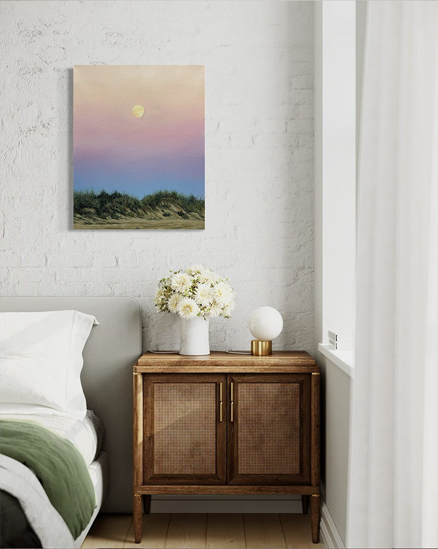 Wooden side table with flowers next to a bed with a painting above on a white wall.