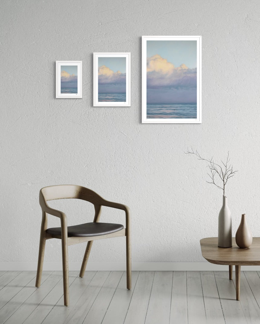 Three framed artworks of a landscape with clouds on a wall above a chair and table.