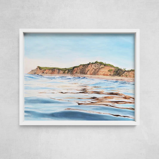 A framed fine art paper print depicting a coastal scene with water in the foreground and cliffs in the background, set against a white wall.