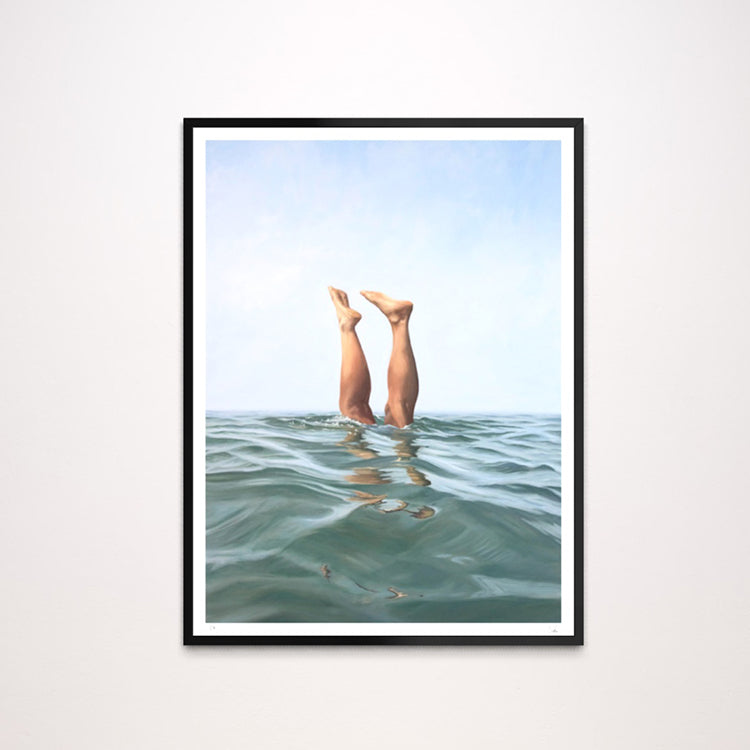 A framed art print featuring a surreal photograph of a person's legs reaching up from the ocean water towards a pale blue sky, presented in a black frame with a white mat against a light gray wall.