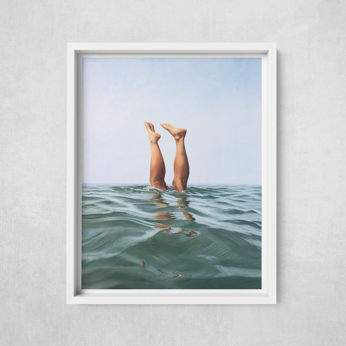A framed art print of two legs emerging from the water, displayed in a white frame against a wall.