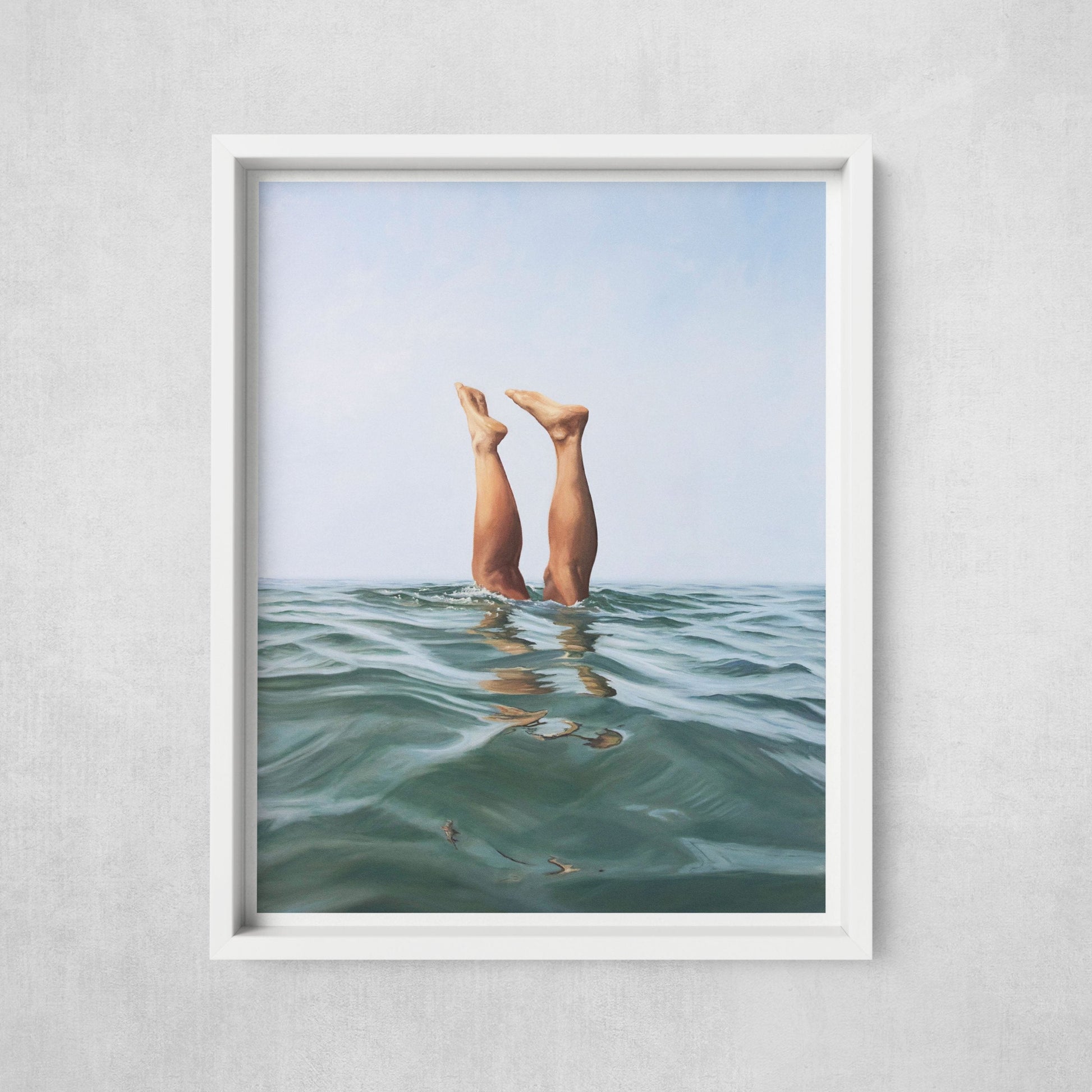 A framed art print of two legs emerging from the water, displayed in a white frame against a wall.