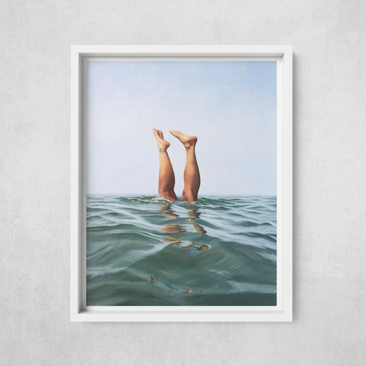 A framed art print of two legs emerging from the water, displayed in a white frame against a wall.