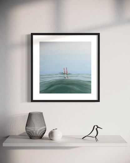 A framed print of 'Double Dipped, I' hanging on a wall above a shelf with a decorative vase and a figure. The artwork depicts two legs emerging from water.