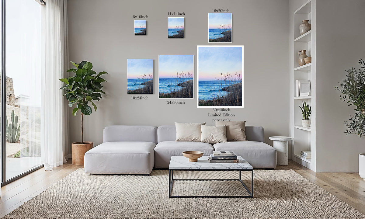 A staged living room with a light gray sectional sofa, coffee table, rug, shelving, and several framed ocean landscape prints mounted on the wall above the couch. The prints show different sizes and one limited edition version, with text labels beneath them.