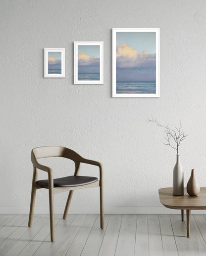 Three framed artworks of a landscape with clouds on a wall above a chair and table.