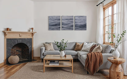 Original ocean triptych wall art above gray sofa in modern coastal living room with fireplace.