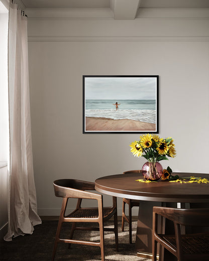 A framed square art print depicting a person running on a sandy beach with ocean waves, displayed on a light gray wall above a wooden dining table with chairs and a sunflower vase.