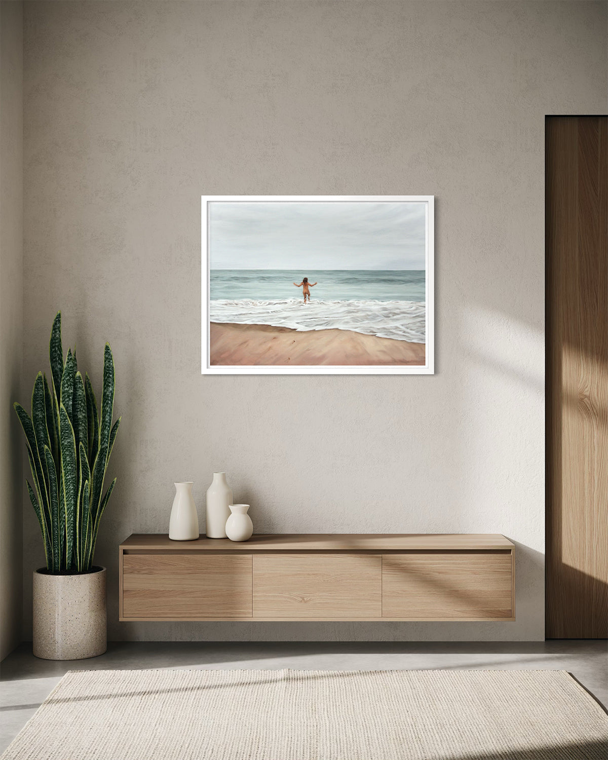 A framed art print depicting a beach scene with a person standing in the ocean, displayed on a textured beige wall above a light wood console table in a modern interior setting.