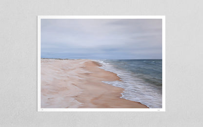 A framed beach landscape art print hanging on a light gray textured wall. The print depicts a sandy beach meeting the ocean under a cloudy sky, with the frame and white border visible against the wall.