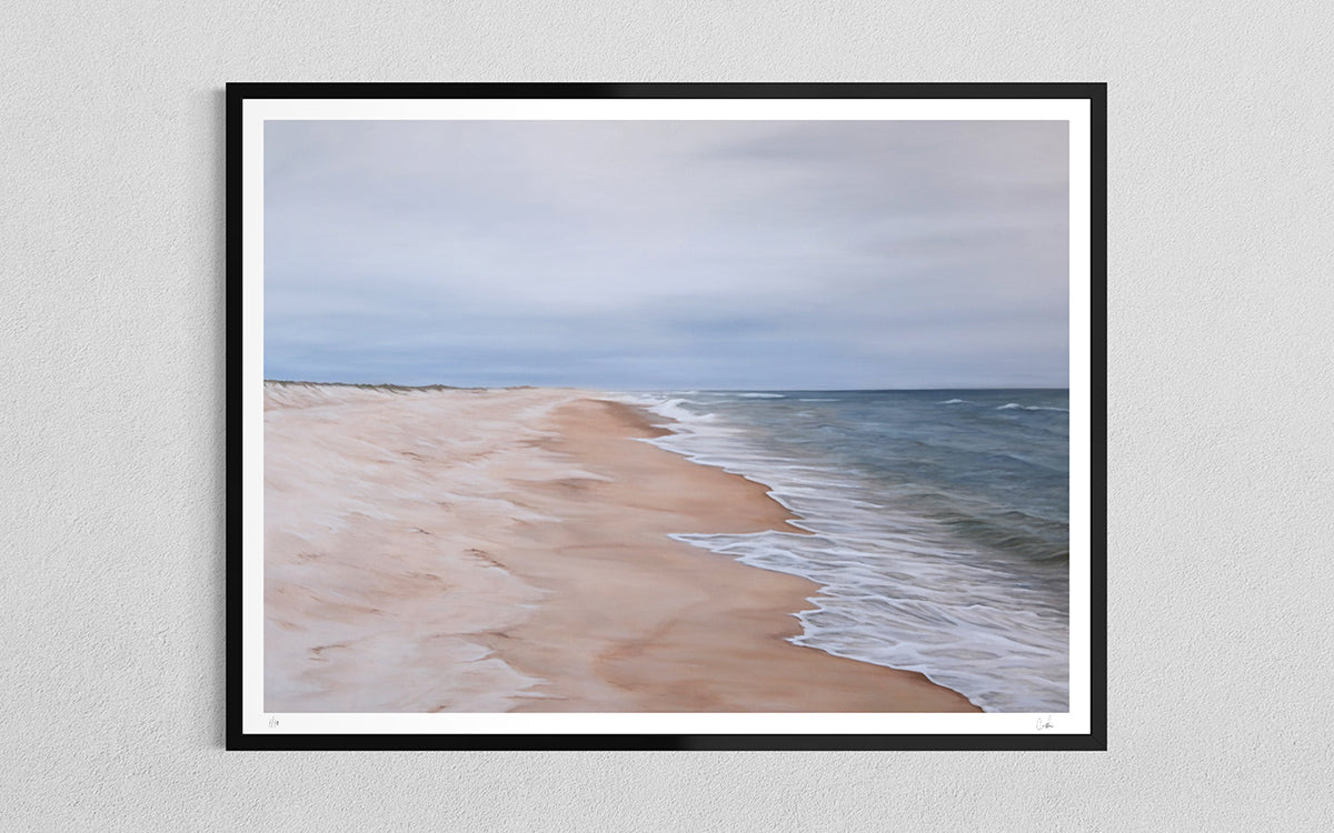 A framed beach landscape print hanging on a light gray textured wall. The artwork shows pale sand, gentle waves, and a cloudy sky, enclosed in a black frame with a white mat.