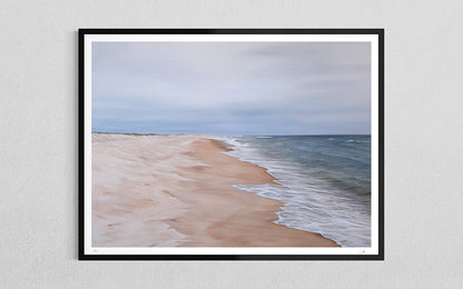 A framed beach landscape print hanging on a light gray textured wall. The artwork shows pale sand, gentle waves, and a cloudy sky, enclosed in a black frame with a white mat.