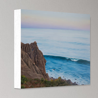 A square canvas wall art print showing a coastal seascape with rocky cliffs, blue ocean water, and a pale pastel sky, photographed against a light gray wall.