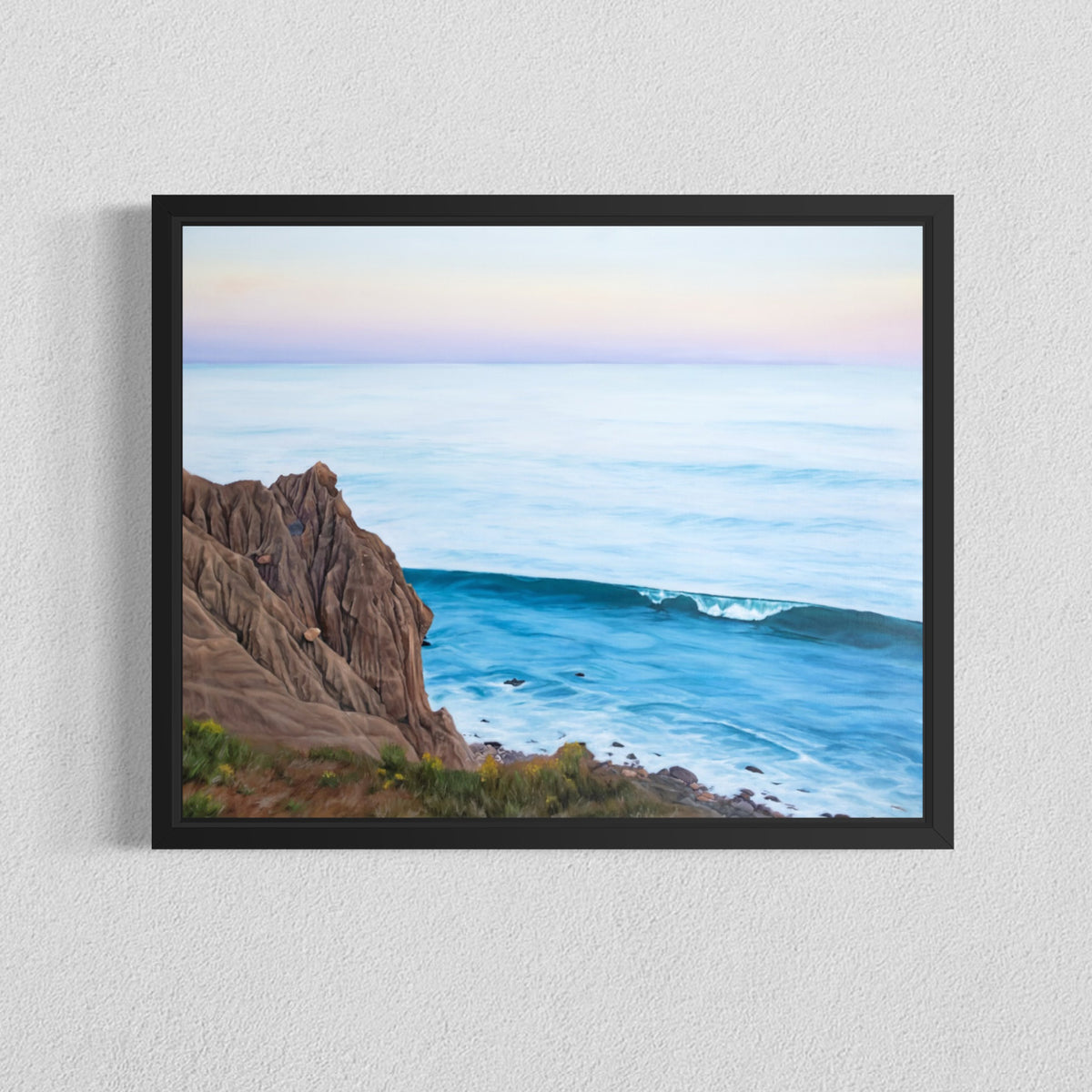 A framed landscape art print depicting a rocky coastline meeting a calm blue ocean under a pale pastel sky, hung on a textured white wall.