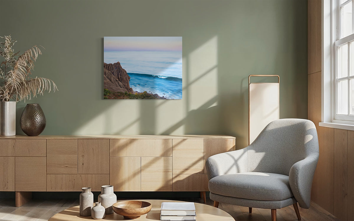 A framed wall art print depicting a coastal cliff and ocean waves, displayed in a modern living room setting with a light wood console, gray armchair, and warm natural light.