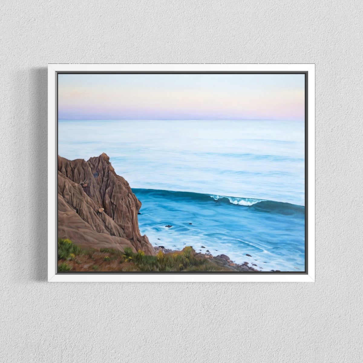 A framed coastal landscape painting hanging on a light gray textured wall. The artwork shows a rocky cliff on the left, blue ocean water, and a pale pastel sky with soft pink and lavender tones.