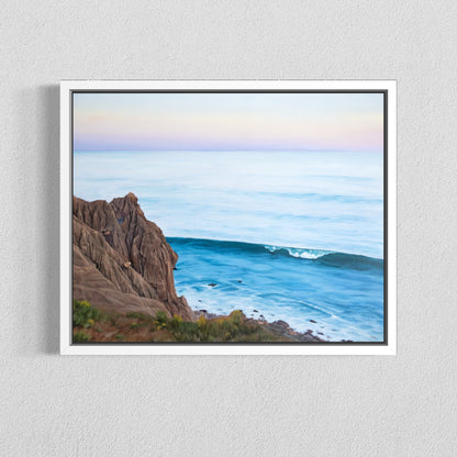 A framed coastal landscape painting hanging on a light gray textured wall. The artwork shows a rocky cliff on the left, blue ocean water, and a pale pastel sky with soft pink and lavender tones.