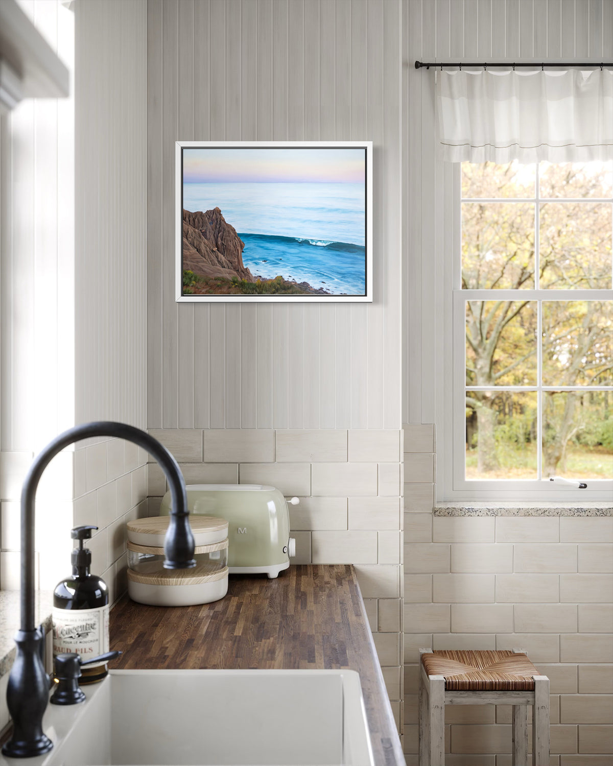 A framed coastal landscape print hangs on a white paneled wall in a bright kitchen. The artwork shows a rocky cliff beside blue ocean water under a pale sky.