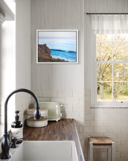 A framed coastal landscape print hangs on a white paneled wall in a bright kitchen. The artwork shows a rocky cliff beside blue ocean water under a pale sky.