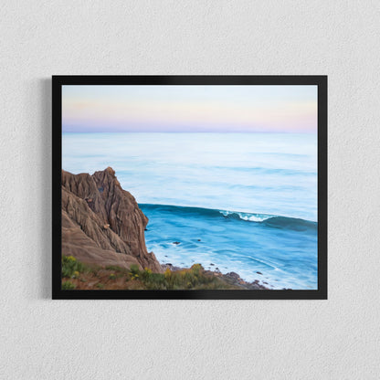 A framed coastal landscape print hanging on a textured white wall. The artwork shows a rocky brown cliff on the left, a blue ocean wave in the foreground, and a pale pastel sky with soft pink and lavender tones.