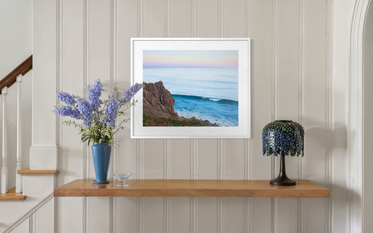 A framed landscape photograph of a rocky coastline and ocean wave, displayed on a light wood console shelf in a home interior with a blue flower vase and a decorative lamp.