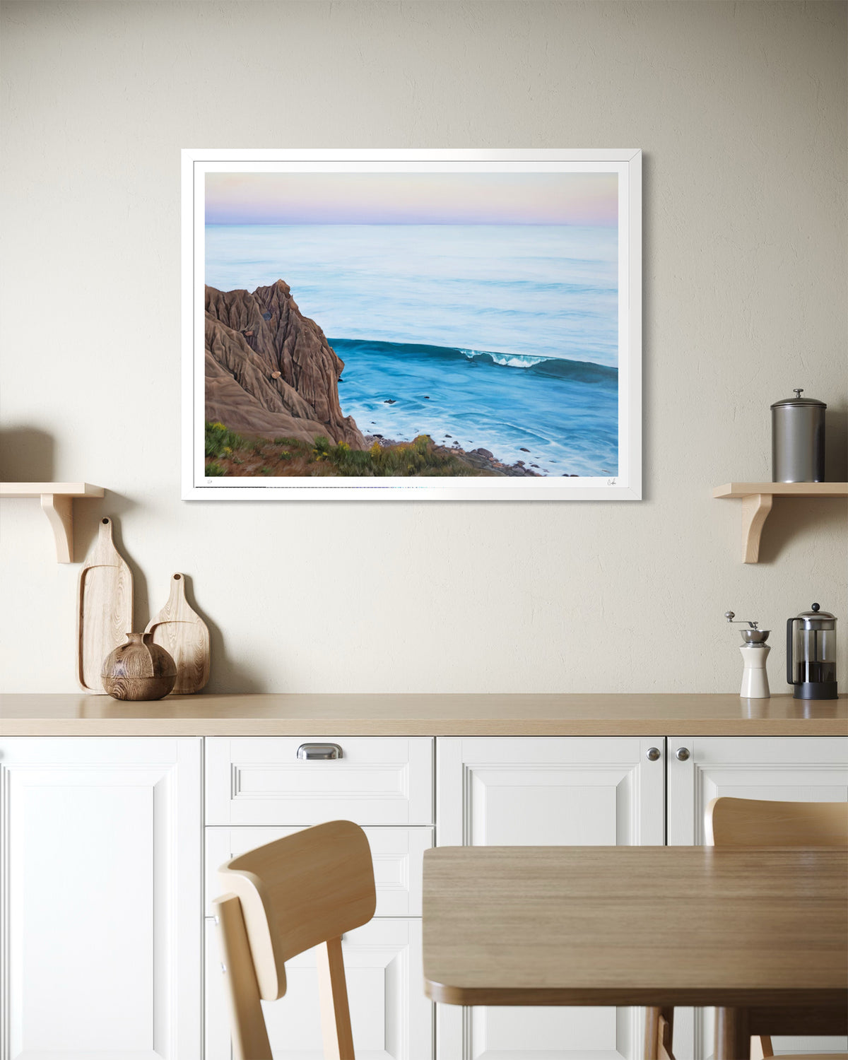 A framed coastal landscape art print hangs on a beige wall above a kitchen counter. The print shows a blue ocean wave with rocky cliffs under a soft pastel sky.