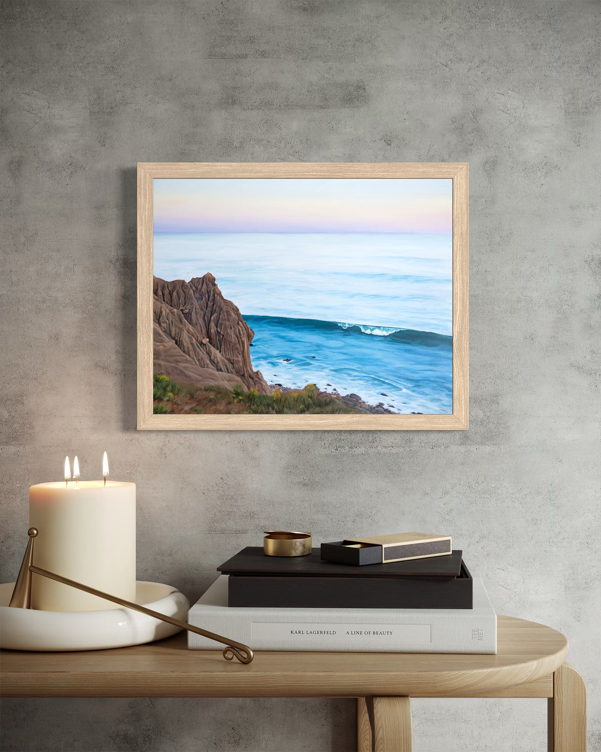 A framed landscape art print of a rocky coastline and ocean waves, styled on a textured gray wall above a wooden table with a candle, books, and a tray.