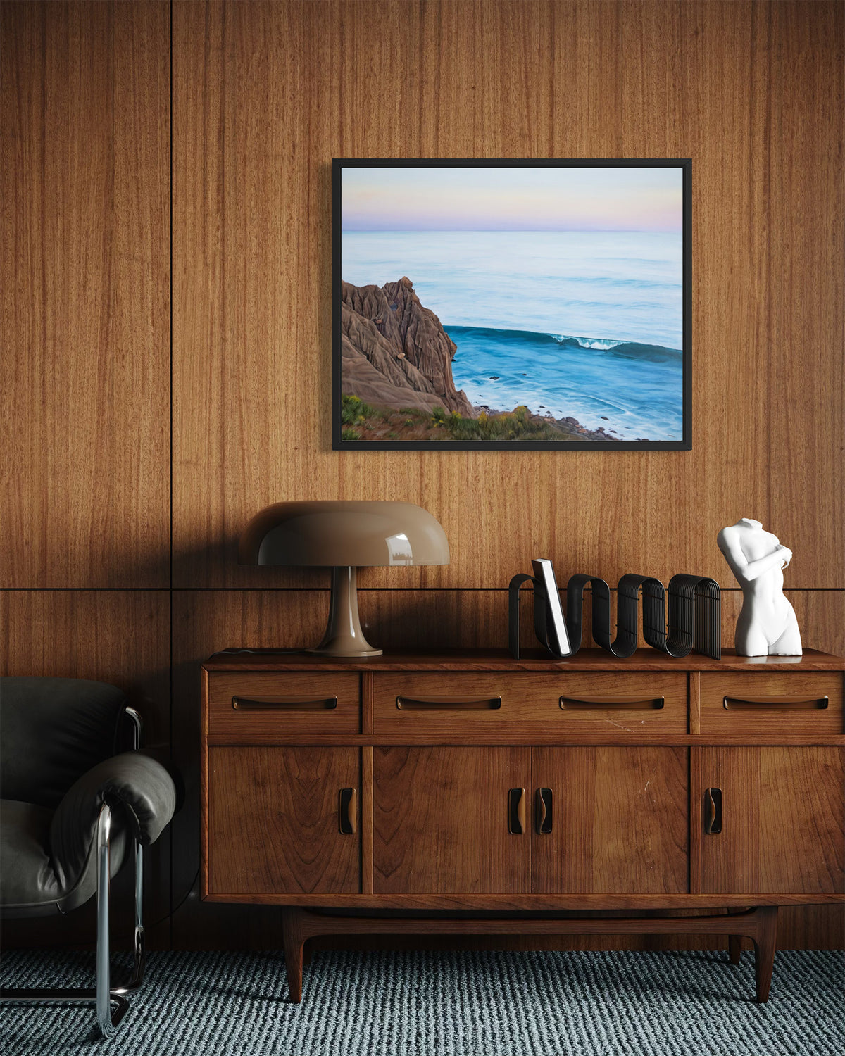 A framed landscape art print of a rocky coastline and ocean wave, displayed on a wood-paneled wall above a mid-century modern wooden credenza in a styled living room.