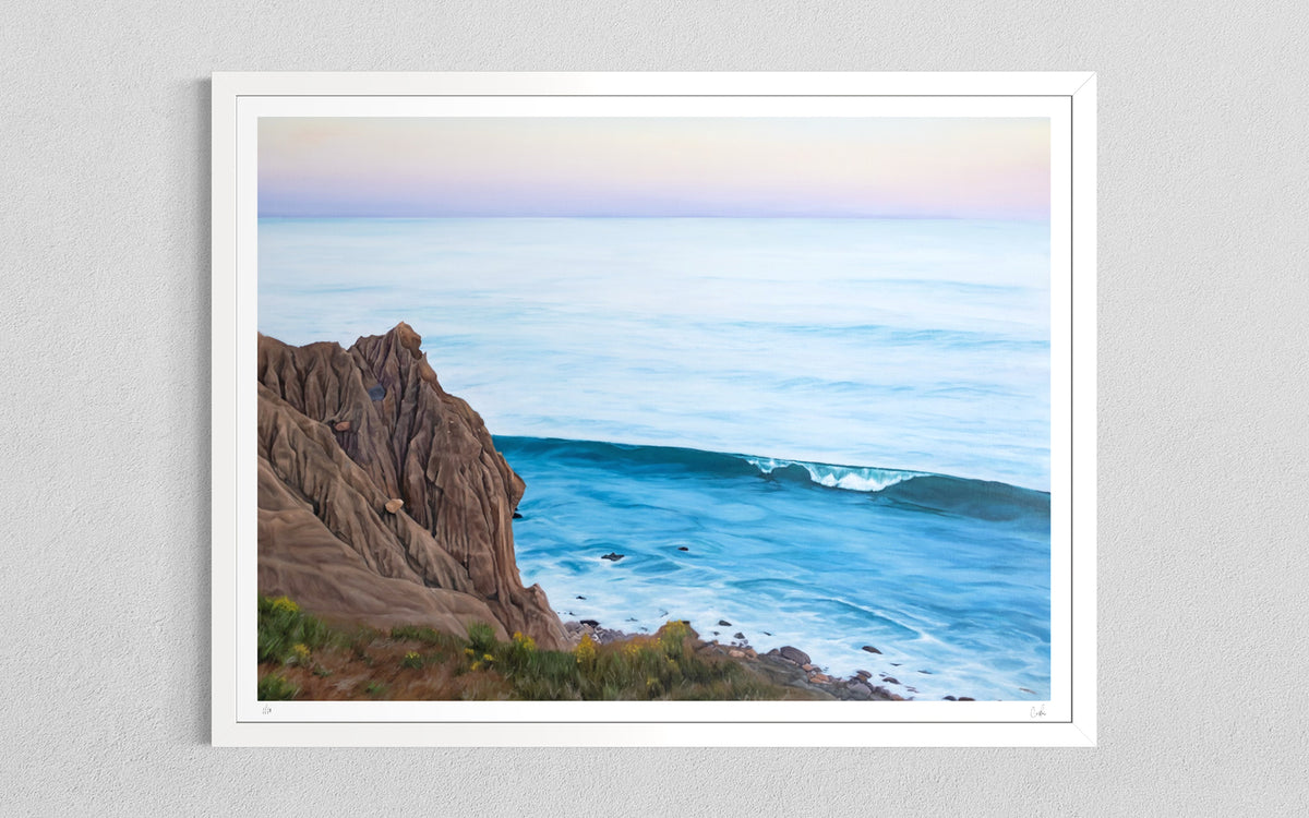 A framed coastal landscape art print hanging on a light gray textured wall. The artwork depicts a rocky brown cliff beside a calm blue ocean with a small breaking wave under a pale pastel sky.
