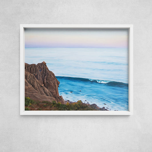 A framed coastal landscape print hanging on a light gray textured wall. The artwork shows blue ocean waves, rocky brown cliffs, and a pale pastel sky, enclosed in a white frame.