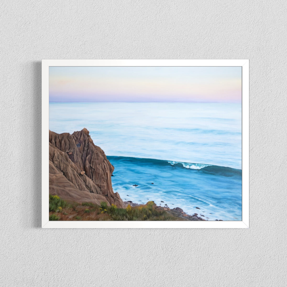 A framed landscape art print depicting a rocky coastline meeting a calm blue ocean under a pale pastel sky, hung on a light gray textured wall.