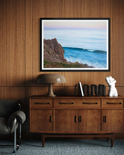 A framed landscape art print depicting a rocky coastline and ocean waves, displayed on a wood-paneled wall above a mid-century modern wooden console table with a lamp and decorative objects.