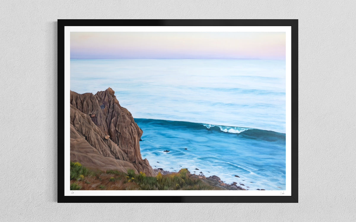 A framed coastal landscape art print featuring a rocky cliff overlooking a blue ocean with a soft pastel sky, mounted on a light gray textured wall.