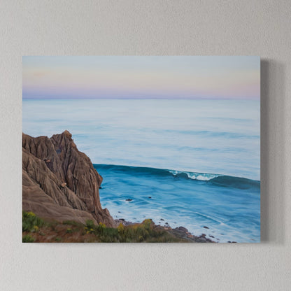 A rectangular canvas print of a coastal seascape hanging on a textured light gray wall. The painting depicts a rocky brown cliff on the left, a blue ocean wave in the center, and a pale pastel sky with soft pink and lavender tones at the top.