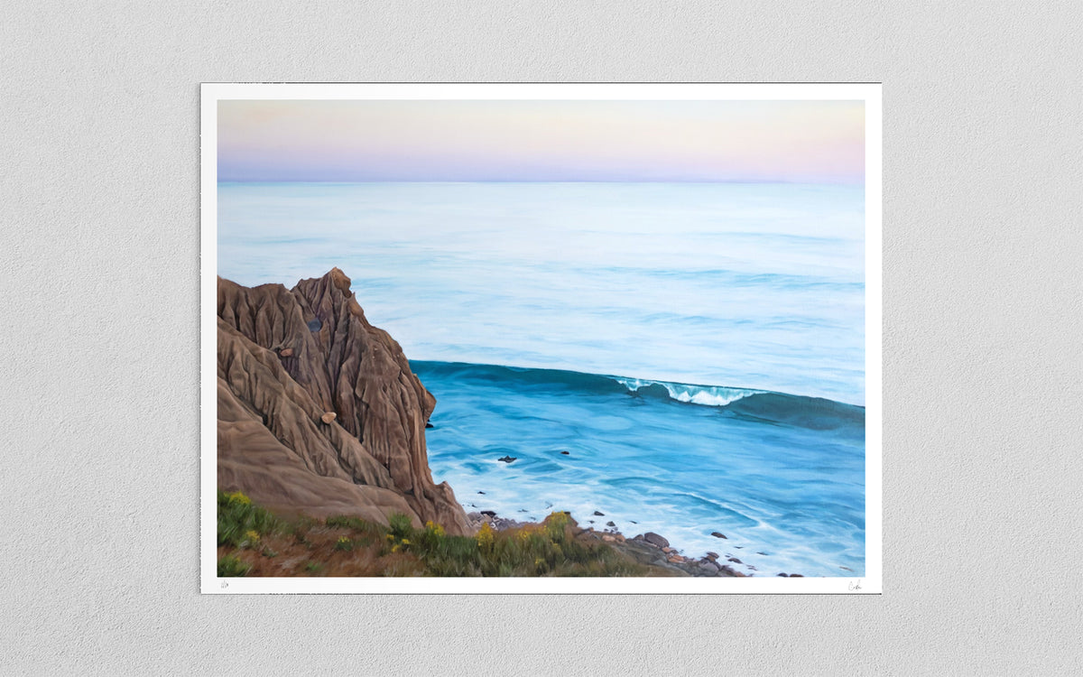 A framed coastal landscape art print hanging on a light gray textured wall. The artwork depicts a rocky brown cliff beside a calm blue ocean with a small breaking wave under a pale pastel sky.