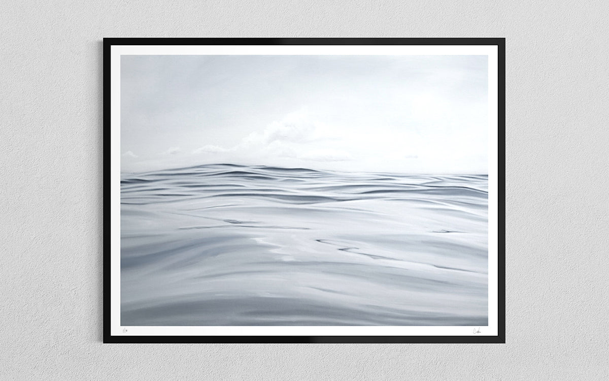 A framed oil painting art print depicting a calm, minimalist ocean with soft gray-blue waves under a pale sky, mounted on a light gray textured wall.