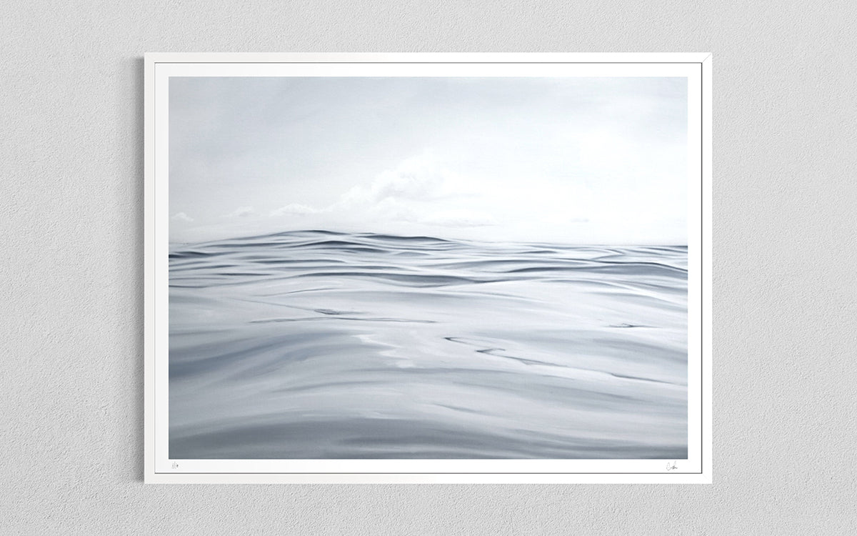 A framed minimalist art print depicting a calm, overcast sea with soft gray and white waves under a cloudy sky, mounted on a light gray textured wall.