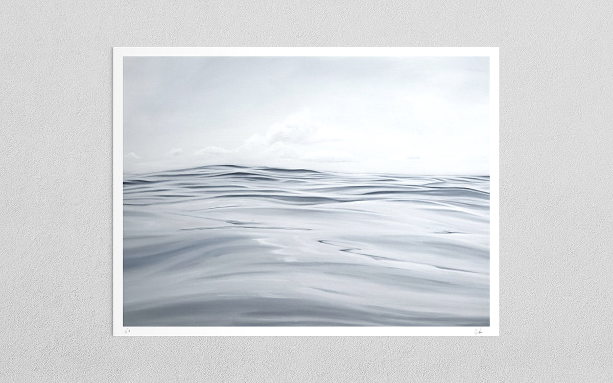 A minimalist art print depicting a calm, overcast sea with soft gray and white waves under a cloudy sky, presented with a white border on a light gray textured wall.