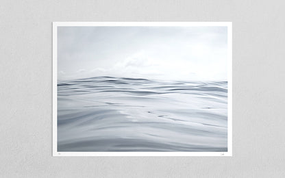 A minimalist art print depicting a calm, overcast sea with soft gray and white waves under a cloudy sky, presented with a white border on a light gray textured wall.