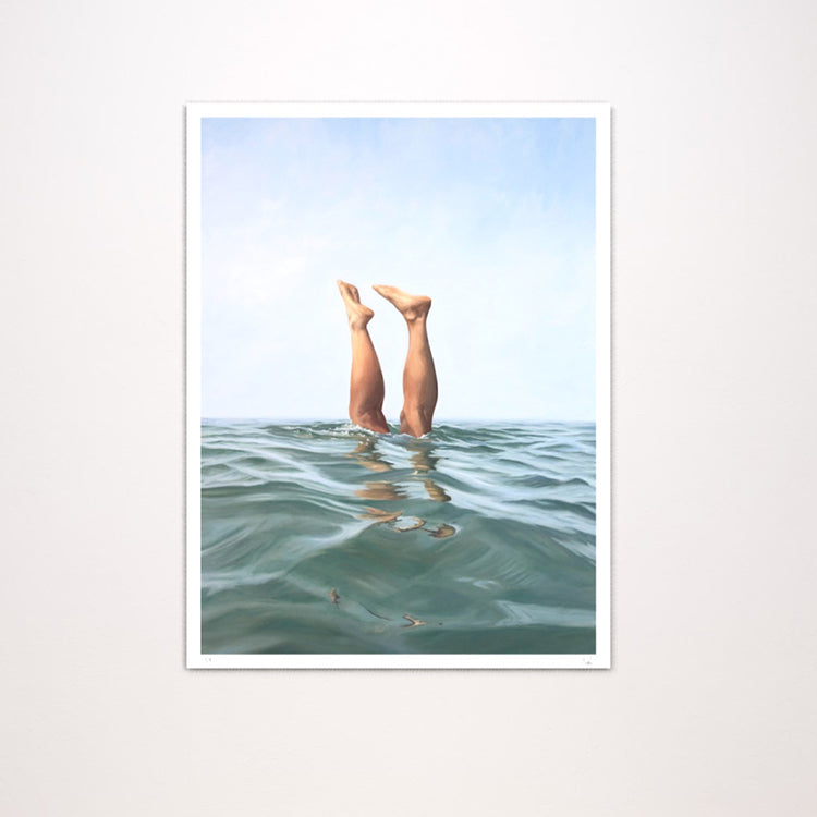 A framed vertical art print featuring a surreal photograph of a person's legs reaching up from calm blue-green water under a pale sky, displayed against a light neutral wall.