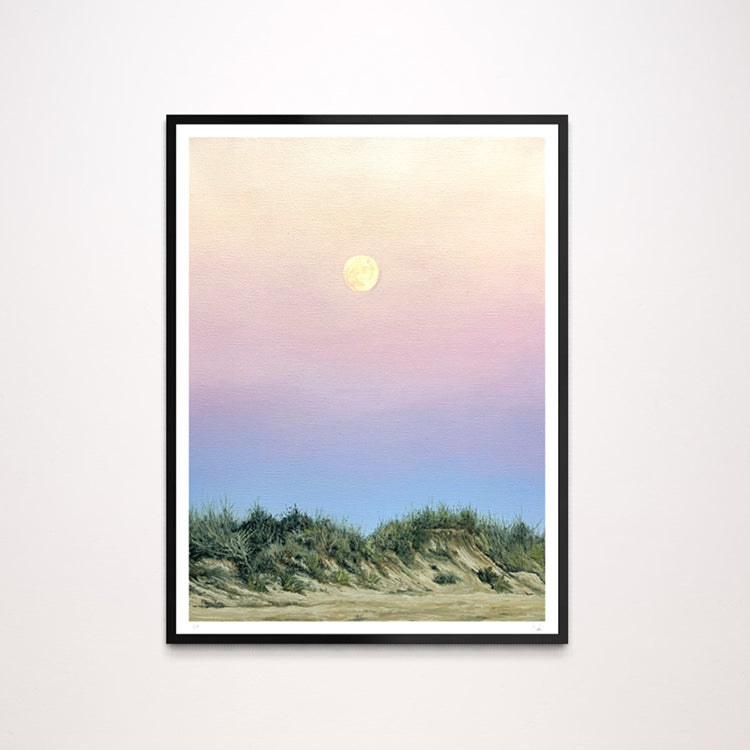A framed landscape art print featuring a pastel sunset sky with a yellow moon over green beach dunes, displayed on a light gray wall.