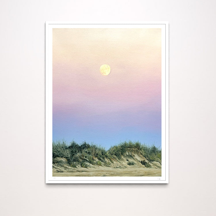 A framed art print depicting a pastel landscape with a yellow moon in a pink and blue sky above dunes, shown against a light gray wall.