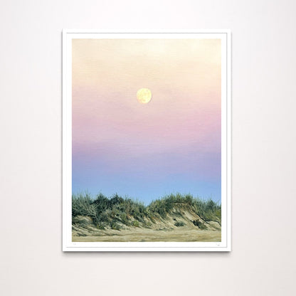 A framed art print depicting a pastel landscape with a yellow moon in a pink and blue sky above dunes, shown against a light gray wall.