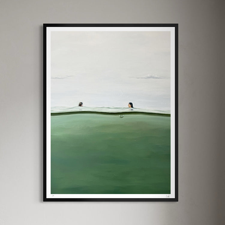 A framed art print depicting a minimalist seascape with two swimmers in a calm green ocean under a pale sky, mounted on a neutral gray wall.