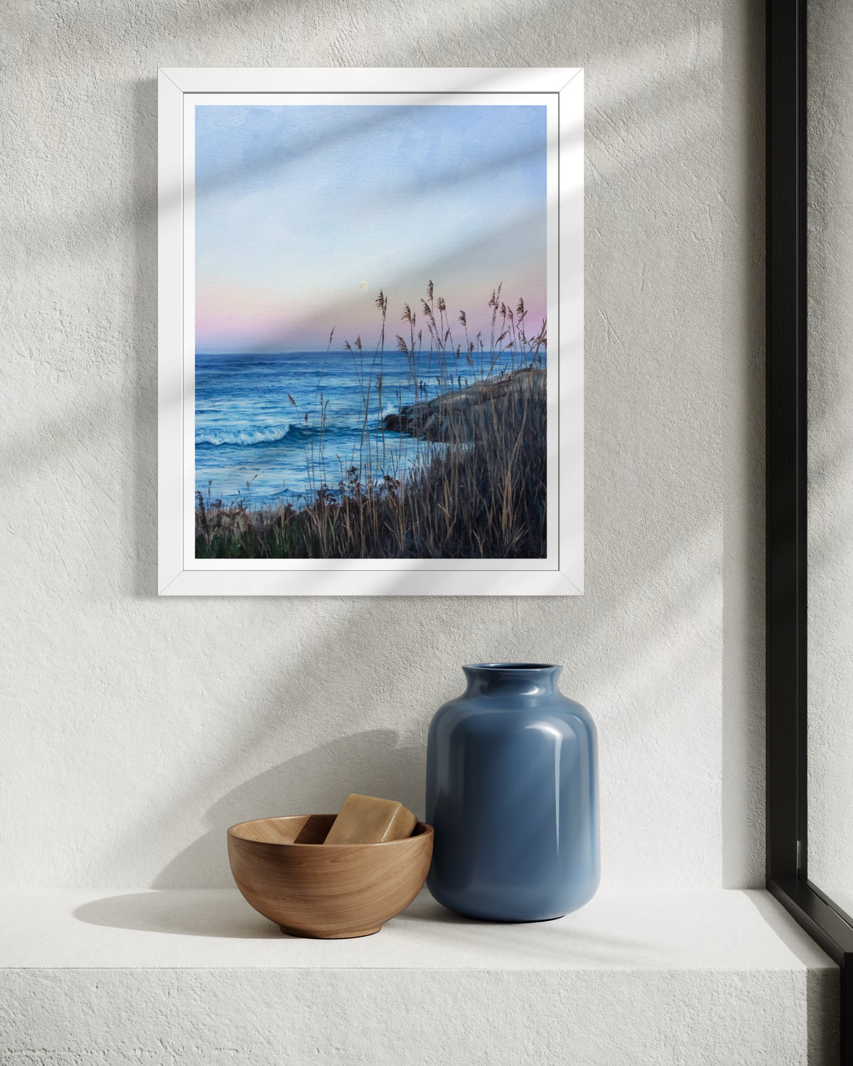 A framed landscape art print of a coastal beach at dusk, styled on a white textured wall above a shelf with a blue vase and a wooden bowl.