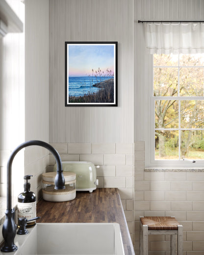 A framed coastal landscape art print hangs on a white paneled wall in a bright kitchen setting with a sink, faucet, toaster, and window.