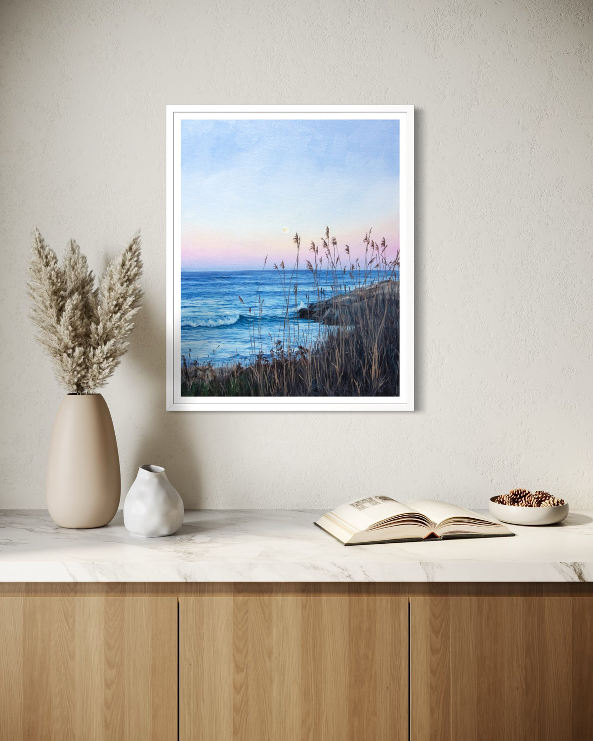 A framed coastal landscape art print hangs on a beige wall above a marble countertop with a vase, small ceramic pot, open book, and bowl in a styled interior setting.